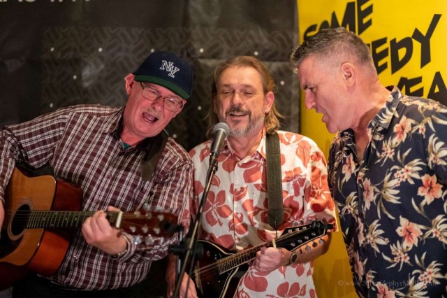 three guys round a mic. One wearing check shirt and baseball cap, other two wearing loud Hawaiian style shirts.
